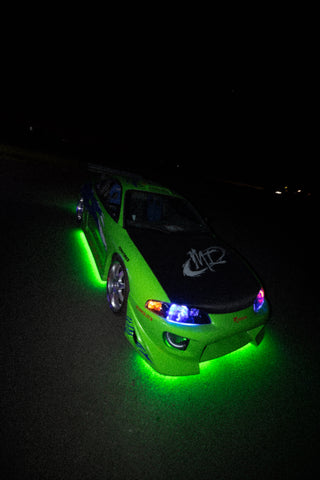 A green and blue Mitsubishi Eclipse with a large rear wing and custom decals, illuminated by green underglow lights.