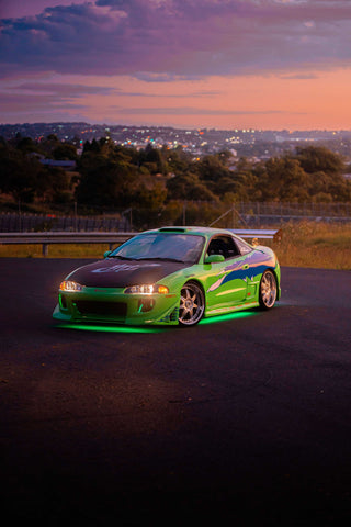 A green and blue Mitsubishi Eclipse with a large rear wing and custom decals, illuminated by green underglow lights.